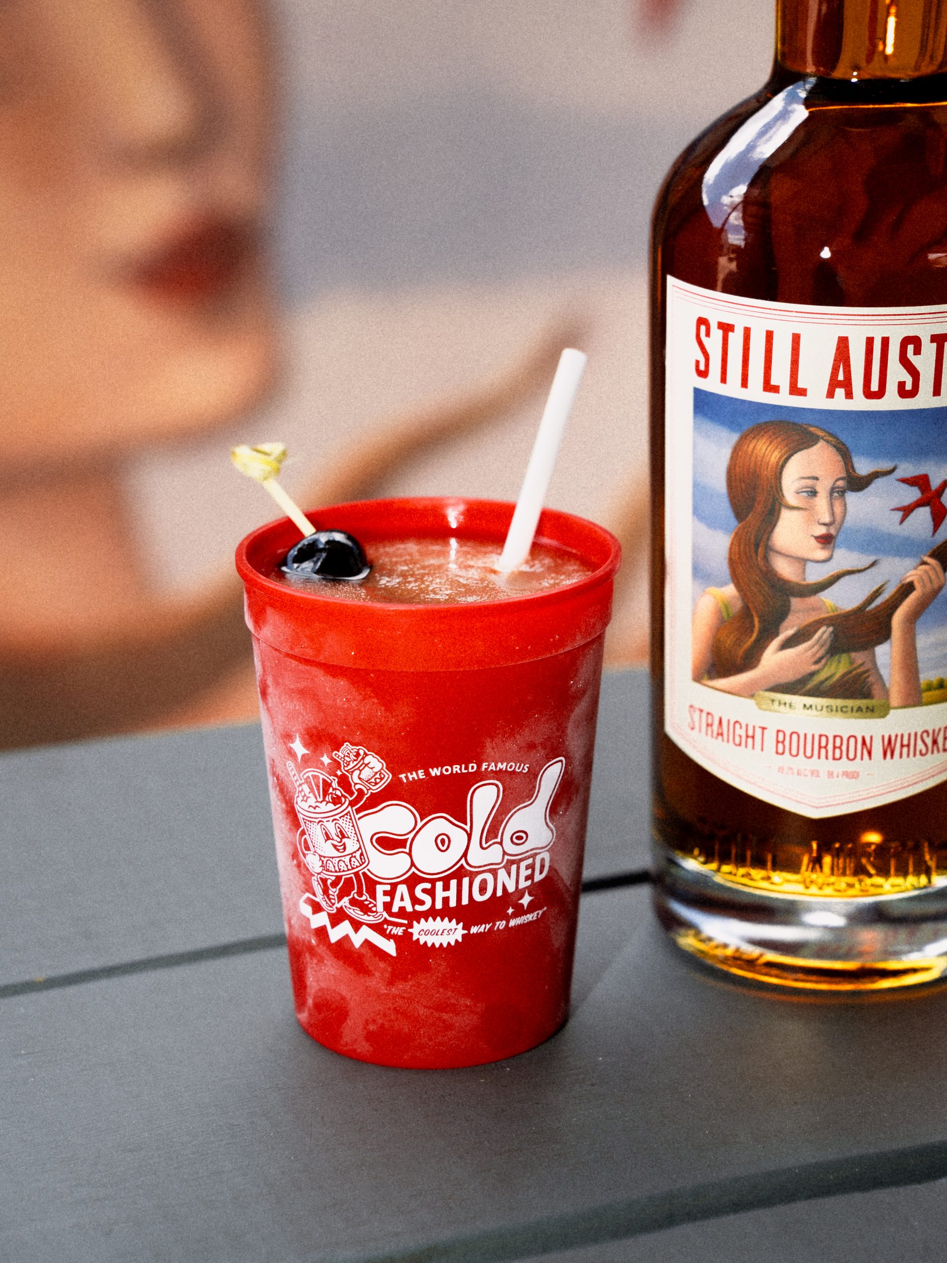 Still Austin Cold Fashioned in red stadium cup beside The Musician straight bourbon whiskey bottle