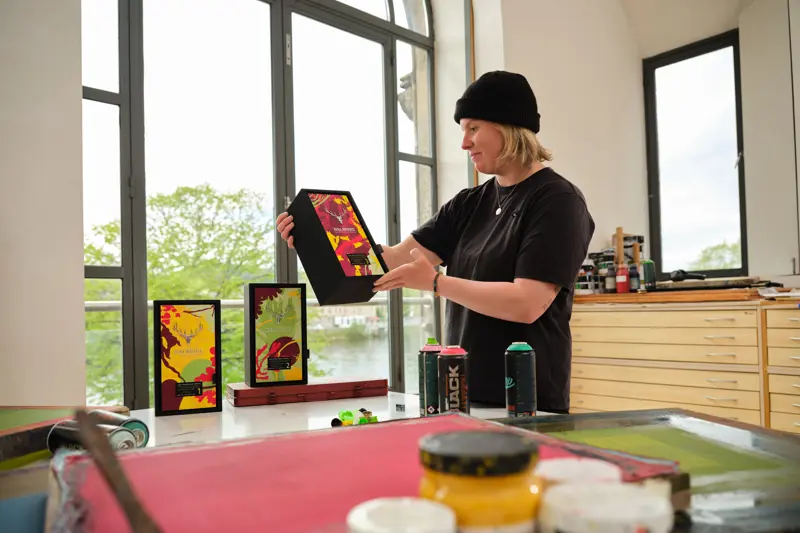 Scottish street artist KMG in her studio holding The Dalmore Portfolio Series 2026 gift boxes, with spray paint cans and screen printing tools