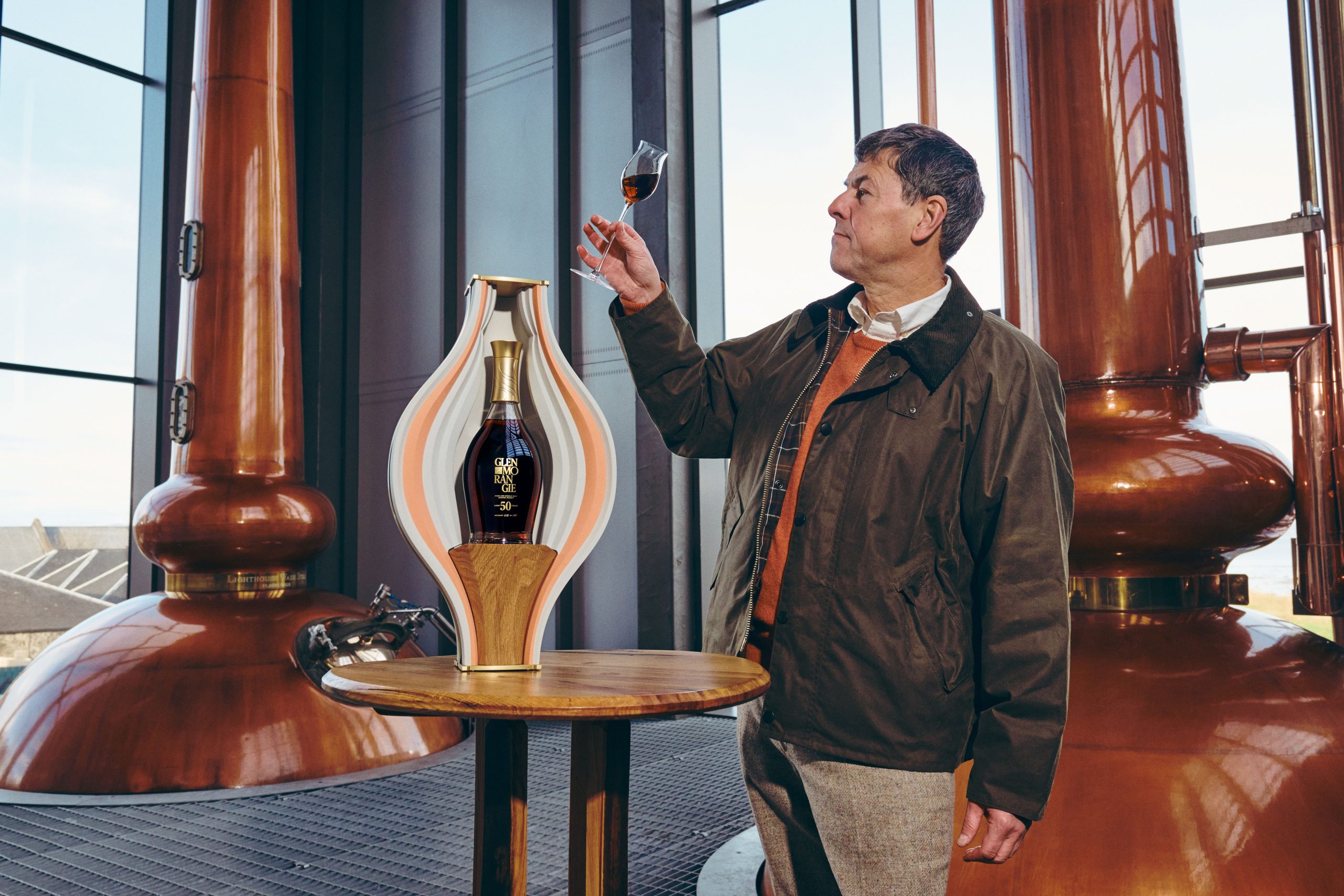 Glenmorangie 50 Years Old in its Peter Pincus porcelain vessel beside the copper stills of Glenmorangie's Lighthouse distillery, as a whisky maker holds a nosing glass to the light