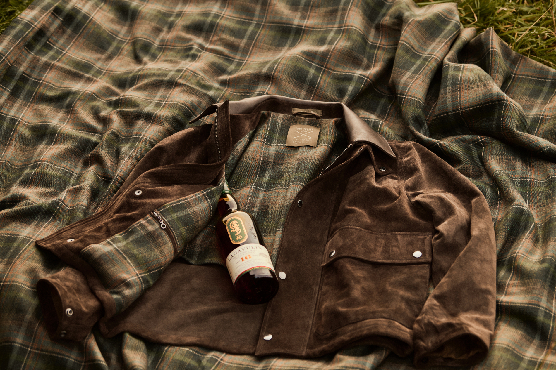 Todd Snyder Lagavulin suede Stable Jacket opened to show its Lagavulin Islay Tartan lining, with a bottle of Lagavulin 16, on a tartan blanket