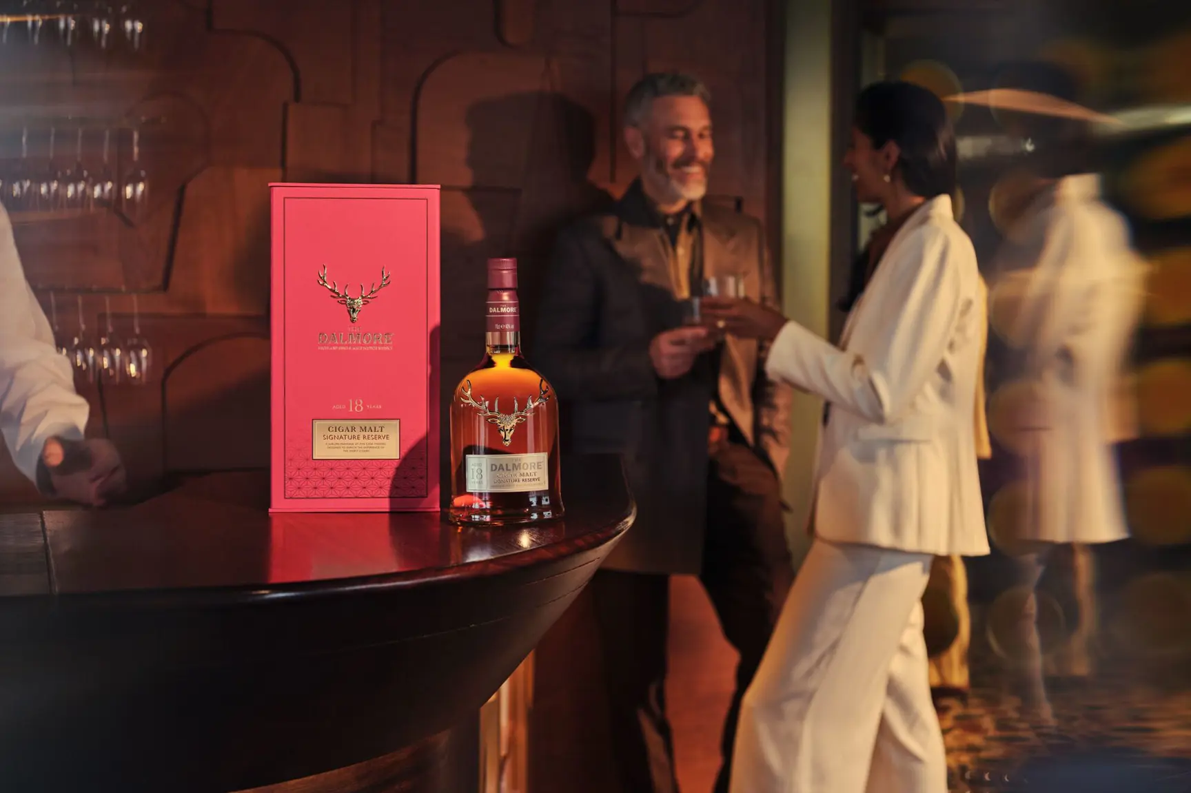 The Dalmore Cigar Malt Signature Reserve 18 bottle and red gift box on a bar counter while two guests share a toast